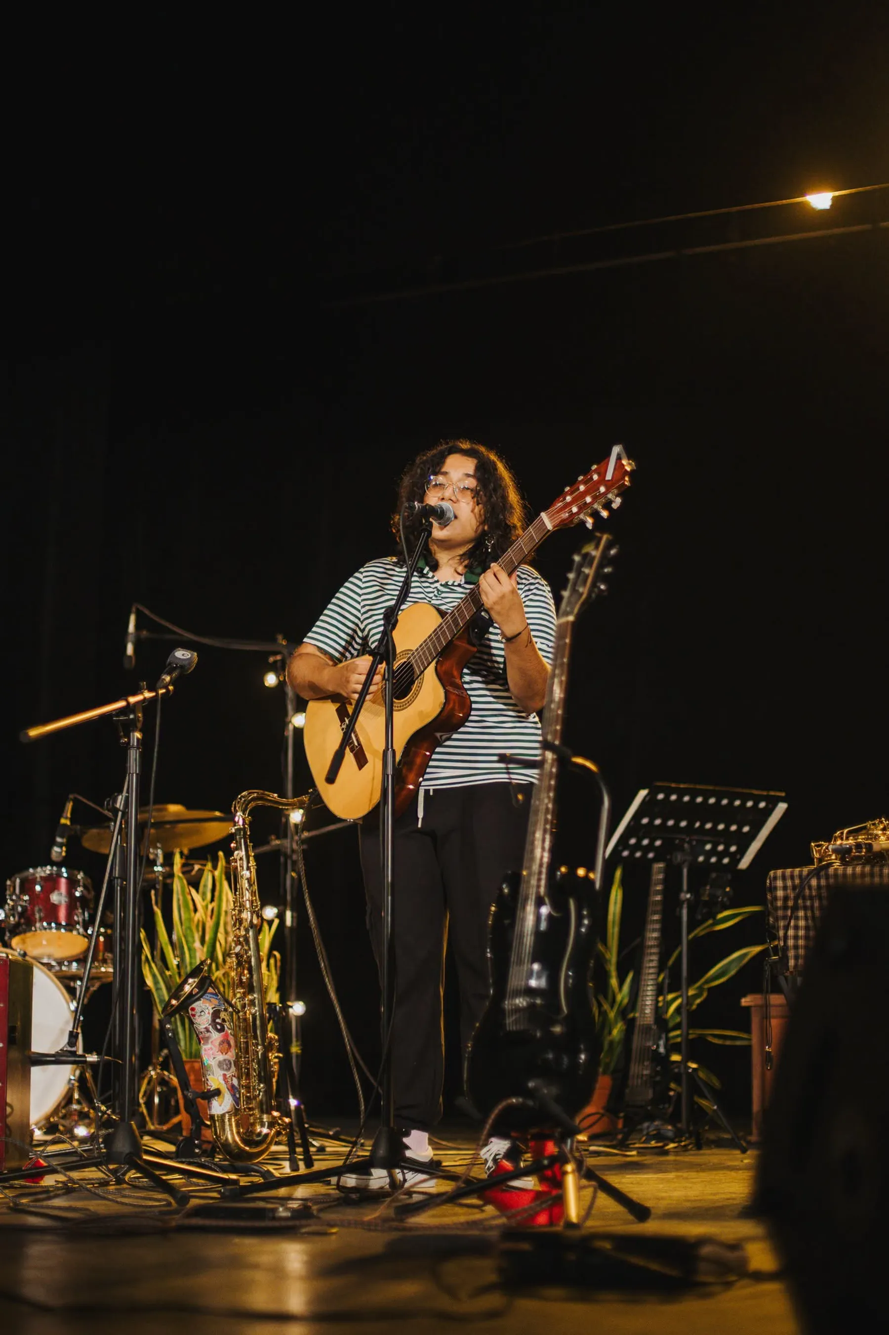 Nelú cantando con su guitarra en un escenario rodeado de plantas, saxofón y batería.