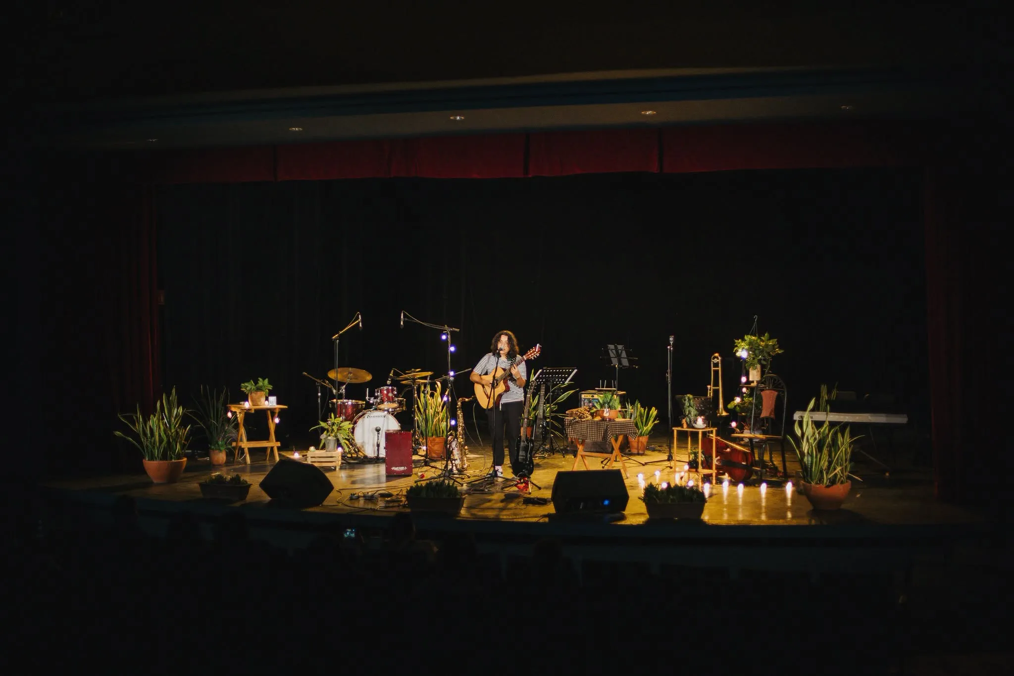 Vista completa del escenario: Nelú al centro con su guitarra, entre plantas, velas y luces cálidas.