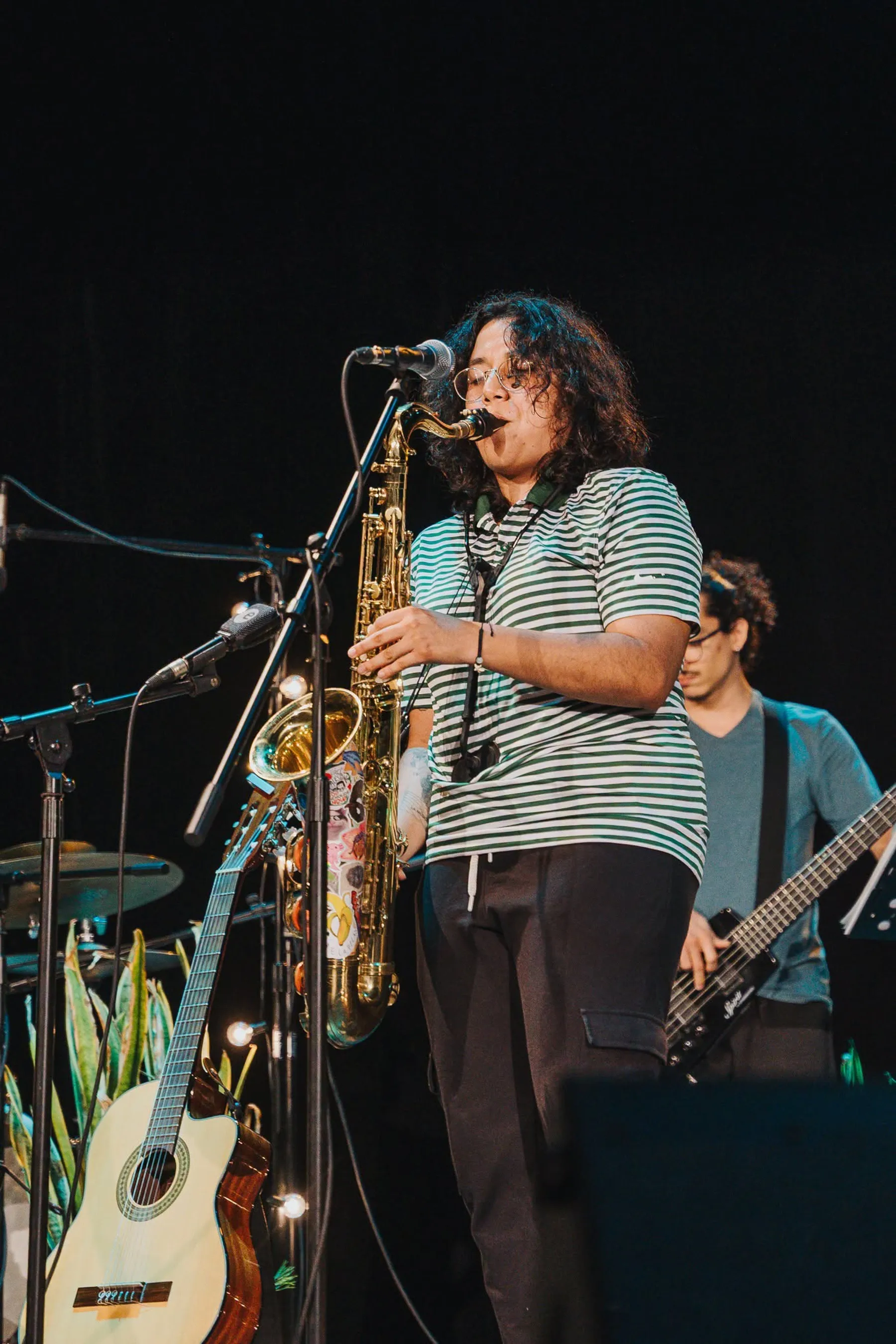 Nelú tocando el saxofón en un escenario oscuro, con camisa de rayas verdes y un bajista al fondo.