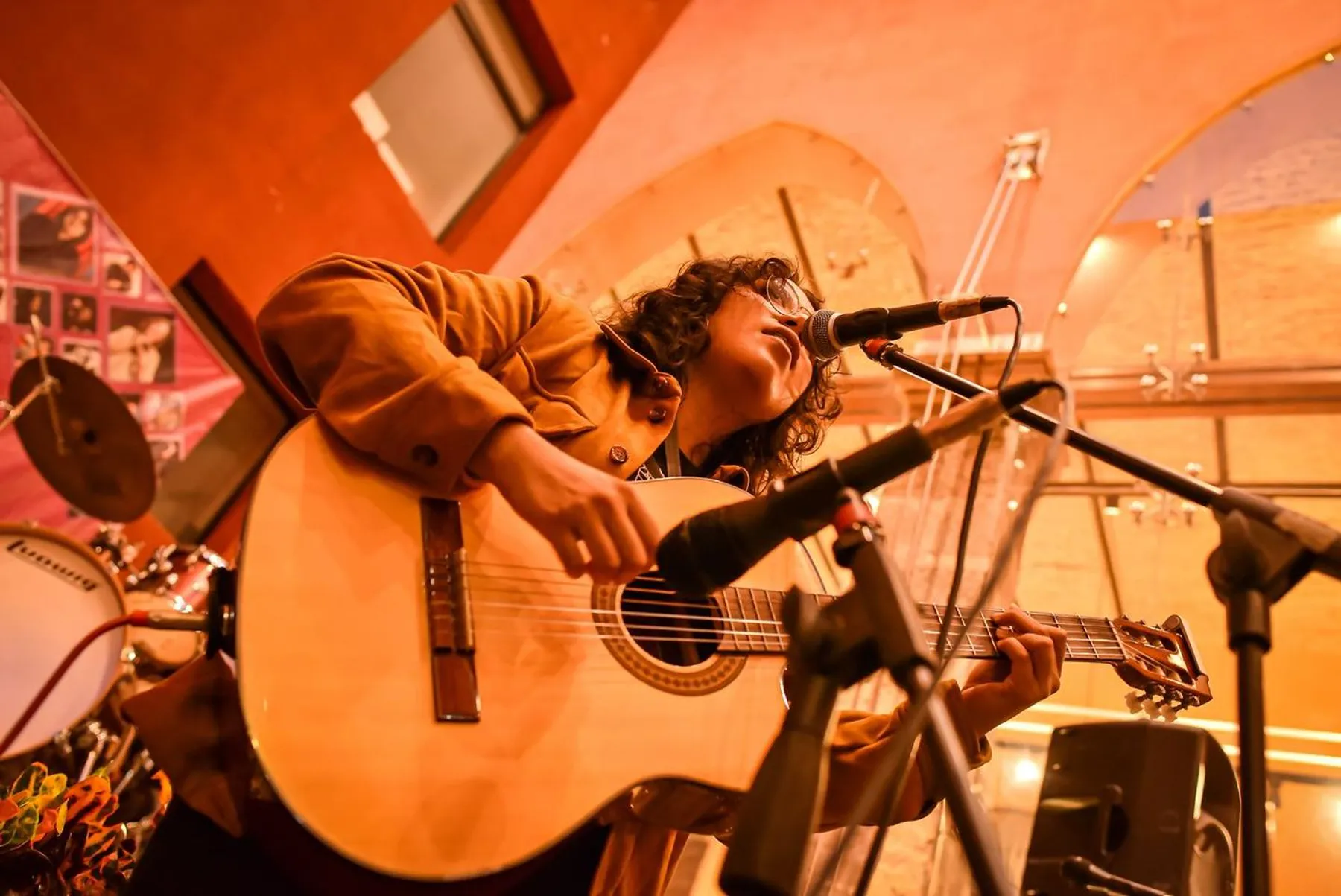 Nelú cantando y tocando la guitarra bajo una luz naranja en un foro, con chaqueta de pana.