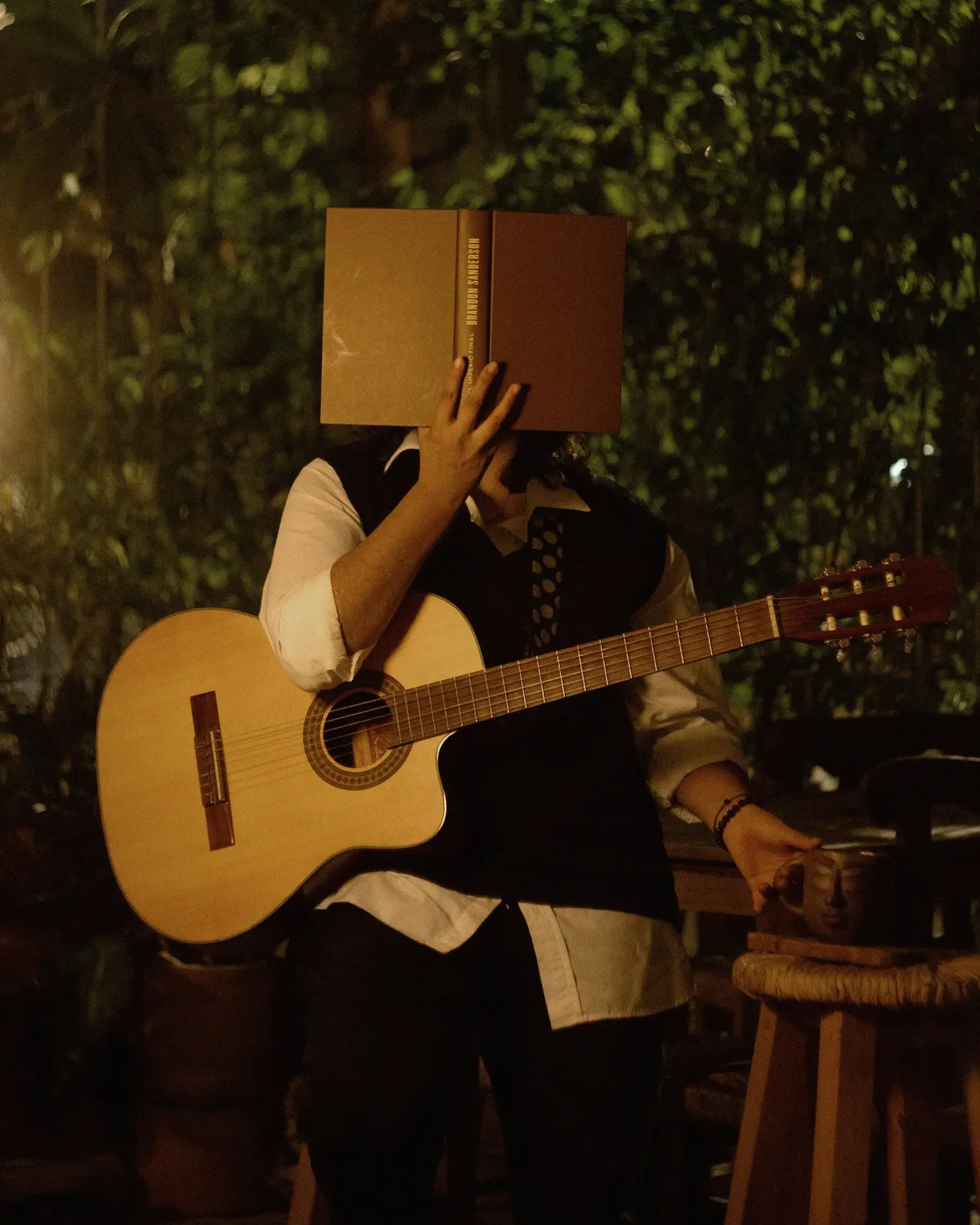 Nelú de pie con una guitarra, cubriendo su rostro con un libro, rodeada de plantas en una noche cálida.