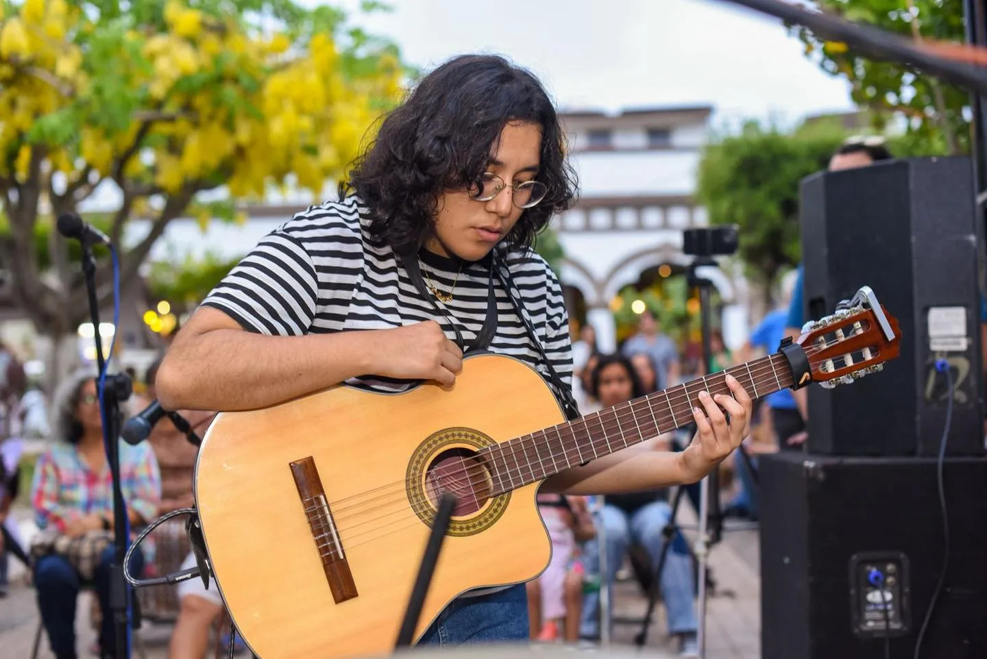 Nelú tocando la guitarra en un concierto al aire libre, con un árbol de flores amarillas y el público detrás.
