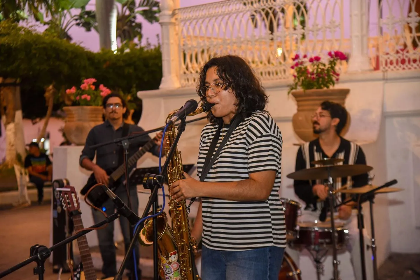 Nelú tocando el saxofón frente al micrófono en un kiosco, acompañada de bajista y baterista.