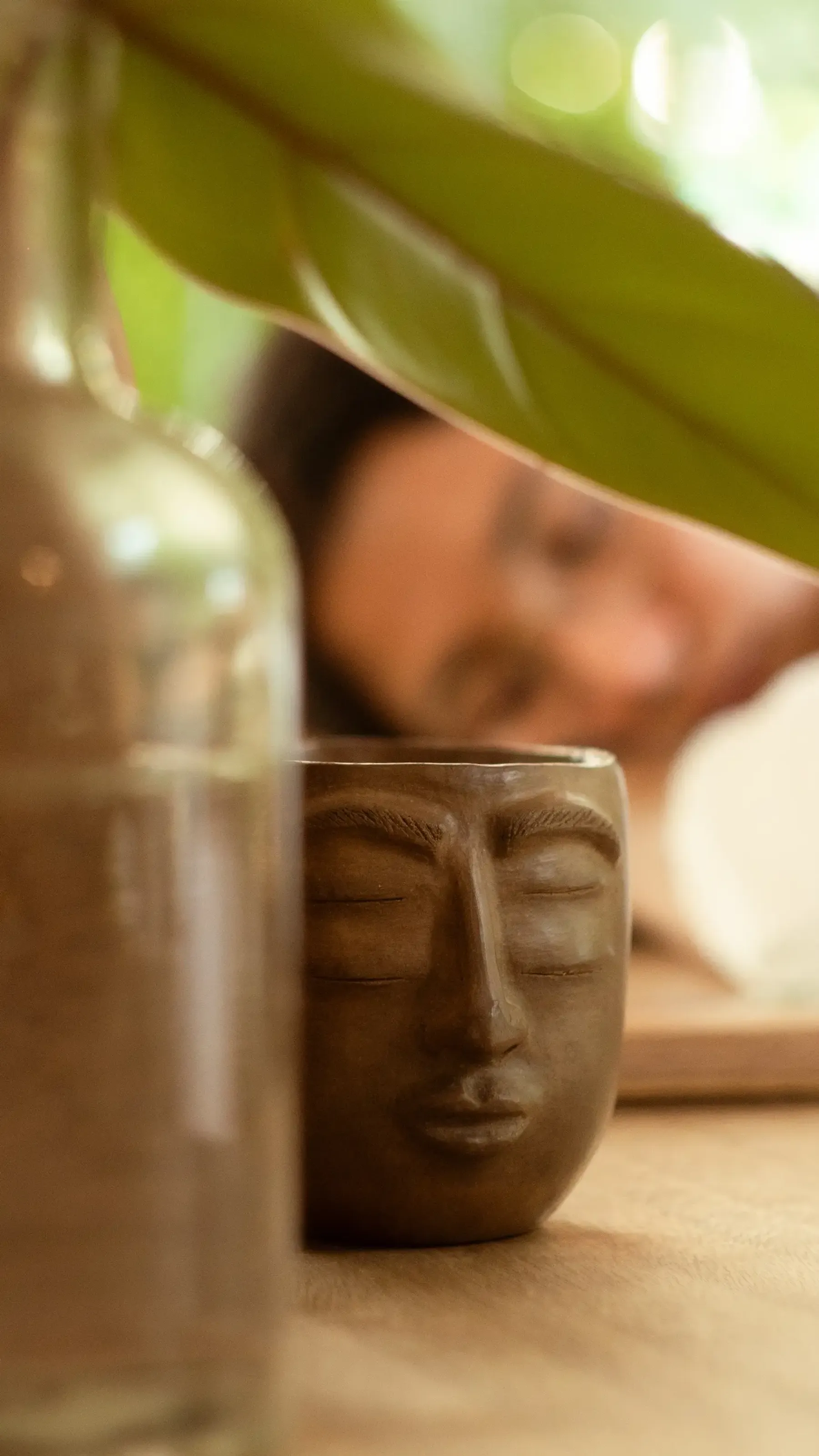 Taza de barro con rostro tallado en primer plano, bajo una hoja verde; Nelú desenfocada al fondo.