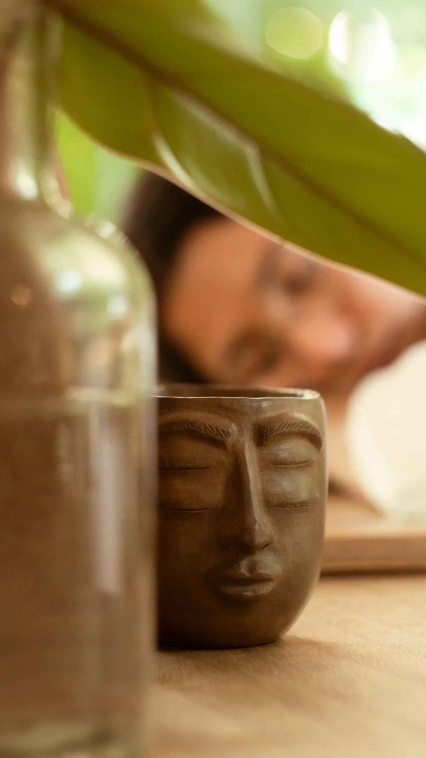 Taza de barro con rostro tallado en primer plano, bajo una hoja verde; Nelú desenfocada al fondo.