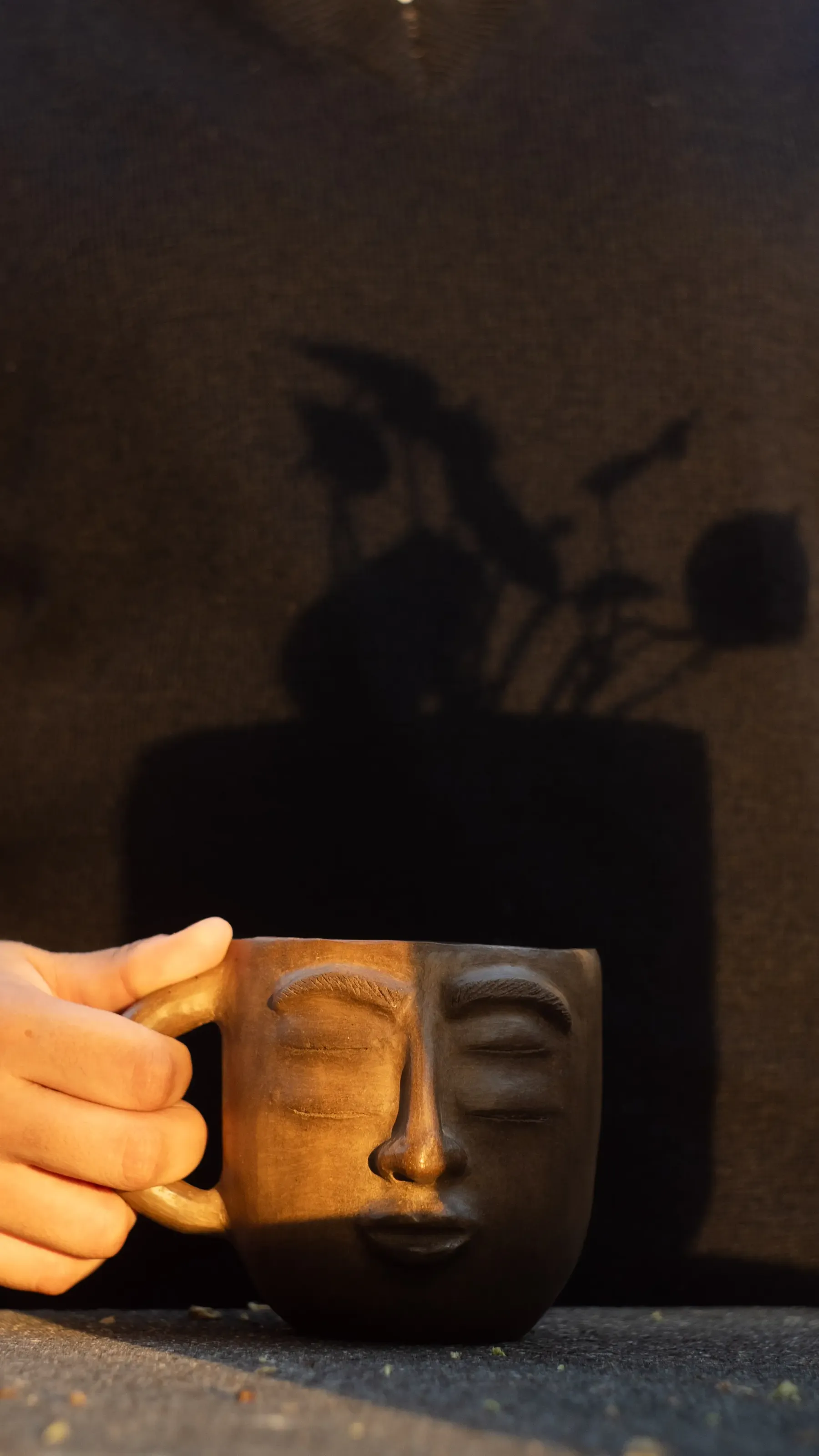 Una mano sostiene una taza de barro con rostro tallado bajo la luz del atardecer, con la sombra de una planta en la pared.